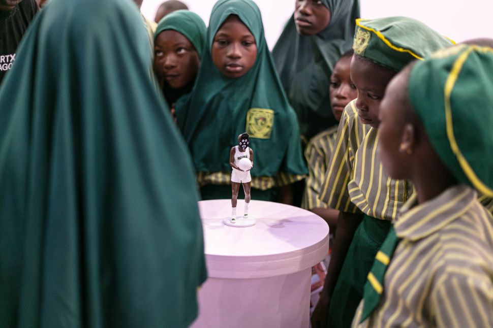 A group of schoolchildren dressed in green uniforms and headscarves gather around a small figurine on a pedestal, focusing intently on the display.