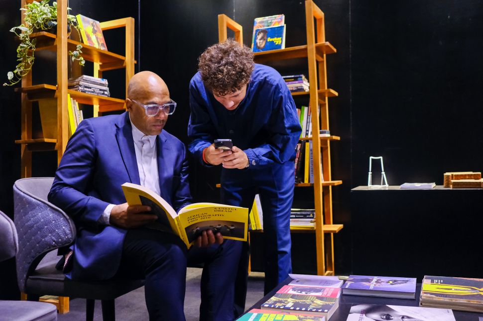 Two men in a library setting look at a book together; one sits and reads while the other stands and takes a photo with his phone, surrounded by shelves of books and dim lighting.