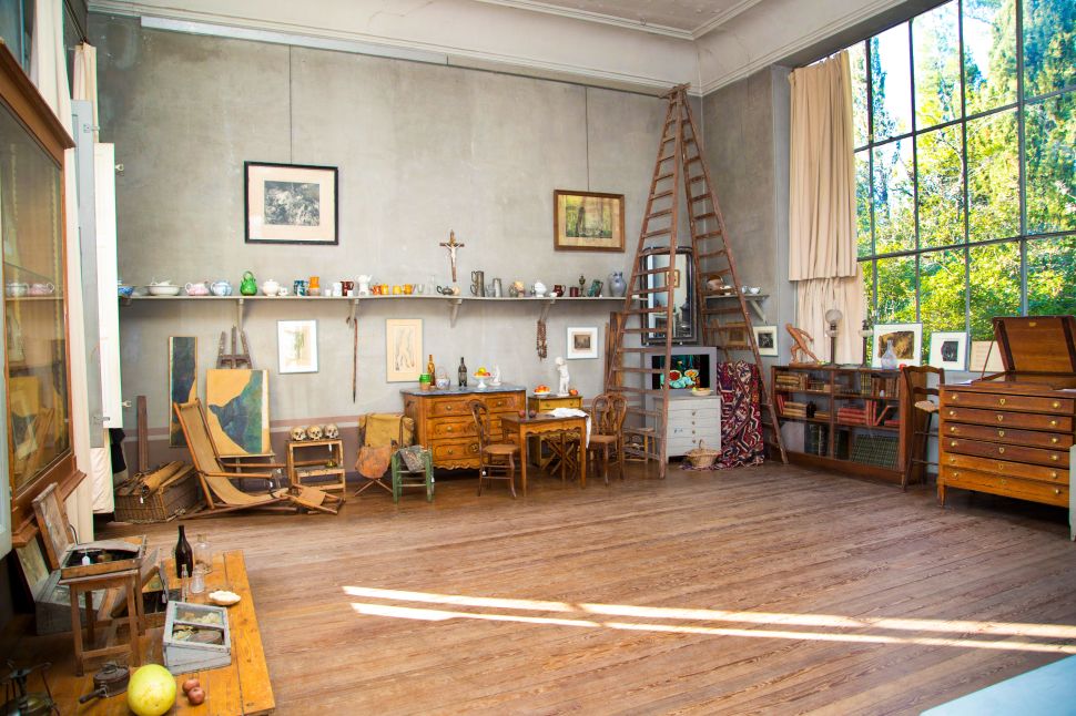 The interior of an artist's studio is shown, with wooden floors, a tall ladder, and large windows allowing natural light to flood the space. The room is filled with shelves of pottery, paintings, furniture, tools, and art supplies.