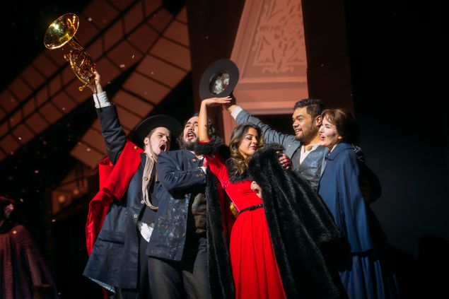 A group of six performers, including a woman in a bright red dress and black fur shawl, celebrate together in dramatic poses, with one man raising a French horn triumphantly. Their joyous expressions and colorful costumes contrast with the dark background, suggesting a climactic or celebratory moment.