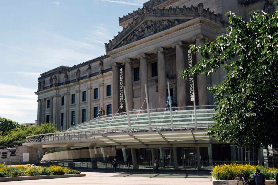 A photograph of the Brooklyn Museum’s grand neoclassical exterior, featuring large columns, banners displaying the museum's name, and a contemporary glass entrance with a landscaped plaza in the foreground, taken on a sunny day.