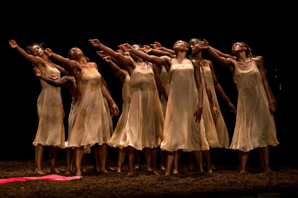 An image of a group of dancers wearing flowing beige dresses performing on a stage covered in soil, with their arms reaching outward in a synchronized motion under dramatic lighting.
