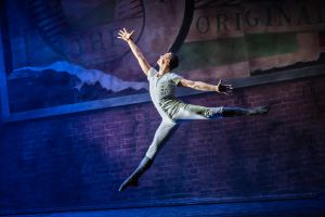 An image of a male ballet dancer in a white costume executing a grand jeté with arms outstretched, leaping across a stage with a brick wall and faded signage in the background.