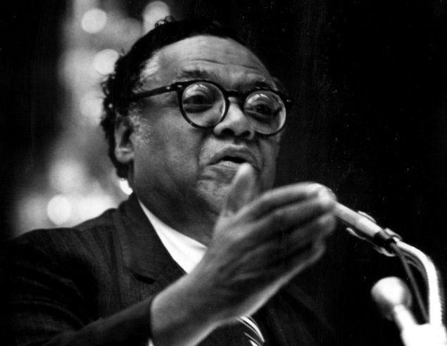 William T. Coleman Jr., wearing glasses and a dark suit, speaks passionately at a podium, gesturing with his hand, exemplifying his role as a leading legal and political figure in American history.