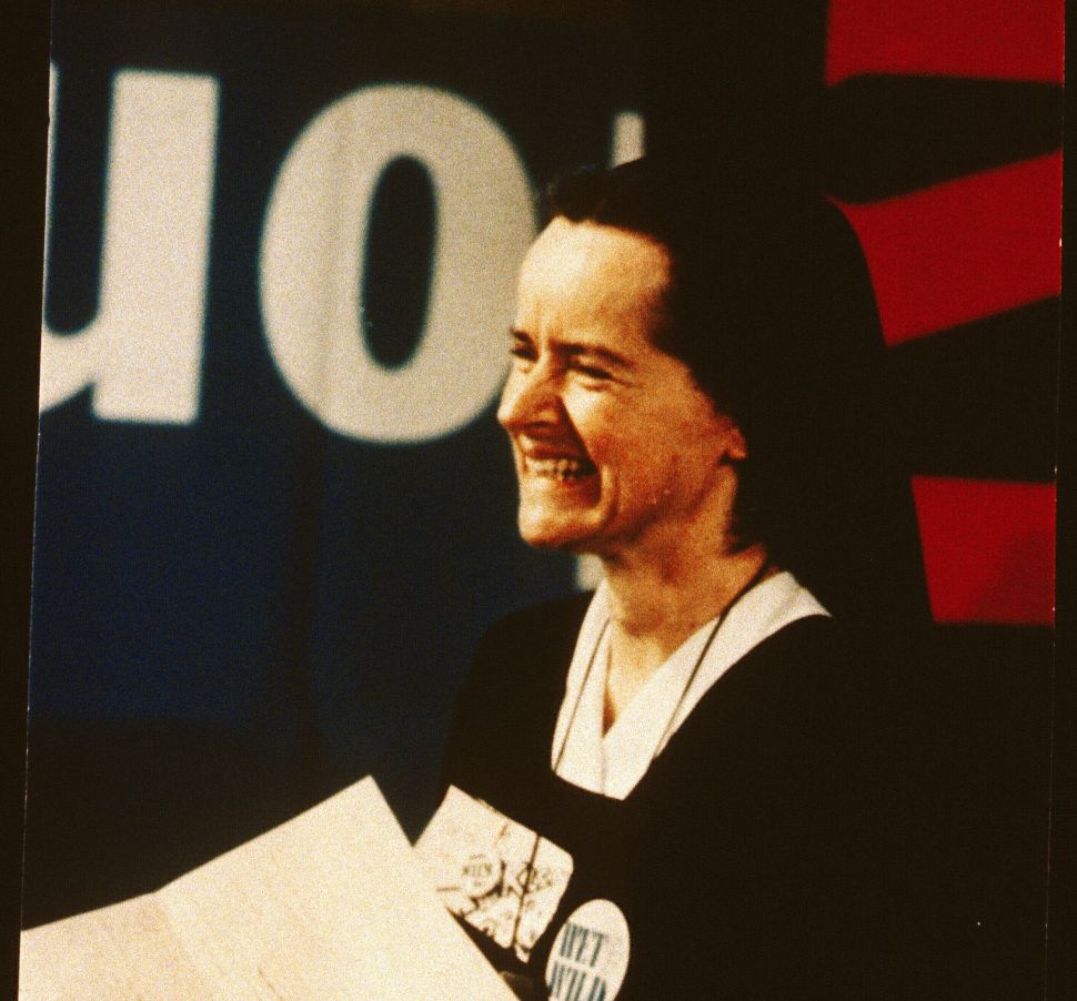 An image of Corita Kent in 1967 shows her smiling broadly while wearing a black nun’s habit and holding sheets of paper, with political buttons pinned to her chest and bold, graphic text banners behind her, reflecting her role as both artist and activist during a time of social upheaval.