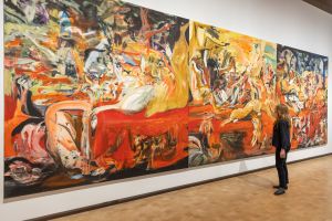 A woman stands in front of a large, wall-sized abstract painting by Cecily Brown composed of vivid swirling colors—primarily red, yellow, and black—with distorted, semi-figurative human and animal forms, inside a contemporary art gallery with wooden floors and white walls.