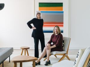 Two professionally dressed women in a modern, minimalist living space with contemporary furniture and abstract artwork in the background. One woman is seated in a wooden lounge chair, while the other stands confidently beside her. The room features neutral tones with a bold, colorful painting as a focal point.