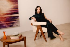 A woman in a long black dress sits confidently in a mid-century modern armchair with cream upholstery and wooden legs, placed in a minimalist gallery space. She faces the camera with a composed expression, next to a round wooden table holding books, a red vase, and a smooth stone. A large abstract woven textile in shades of purple and orange hangs on the wall behind her.