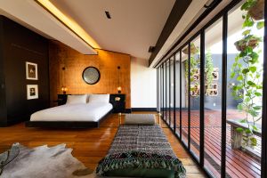 A modern hotel room with warm wood floors, a copper accent wall, and floor-to-ceiling windows opening to a private terrace with hanging plants.