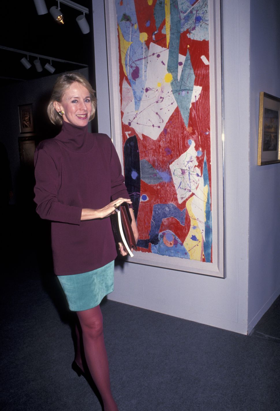 A woman in a plum turtleneck, teal skirt, and red tights smiles while holding books in front of a tall, abstract painting featuring red, blue, white, and yellow shapes in a gallery setting with overhead track lighting