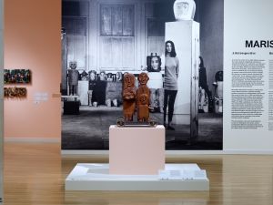 Image of an install view with wood sculptures and image in balck and white of a woman in a studio.