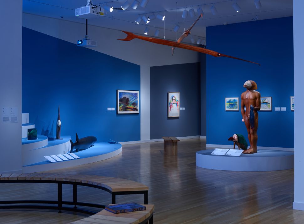 Exhibition space with blue walls and sculptures of sea creatures and human-animal hybrids, including a tall figure with a bird head holding a fish.