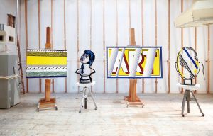 Four Roy Lichtenstein artworks, including paintings and sculptures in his signature pop art style, are displayed on easels and stools inside a bright, unfinished studio space with exposed beams and a worn wooden floor.