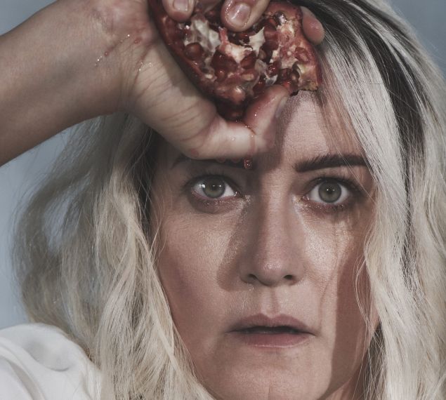 A close-up of a woman with light blonde hair holding a split pomegranate against her forehead, with red juice dripping down, referencing a promotional image for the Metropolitan Opera’s new production of Salome.