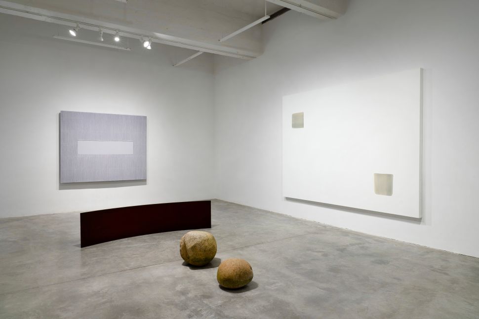 A minimalist gallery installation features two abstract paintings—one with vertical gray lines and a central white rectangle, and the other a large white canvas with two faintly shaded rectangular forms. On the floor, two round stone sculptures and a curved, rust-colored steel sheet add dimensional contrast to the clean, quiet space.