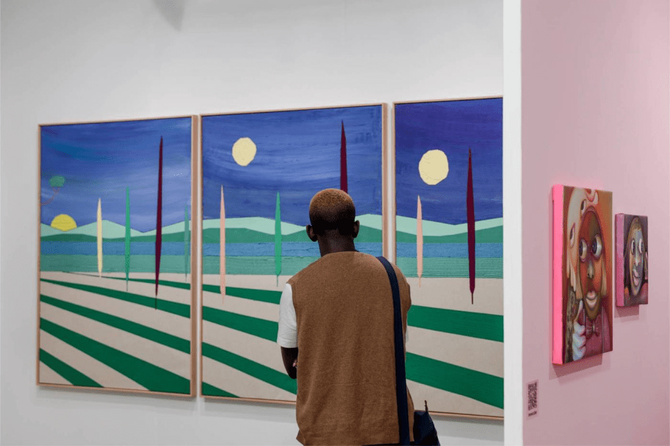 A person with short, bleached hair and a brown vest stands with their back to the camera, observing a vibrant triptych painting of a stylized landscape with trees, hills, and yellow moons against a deep blue sky. Smaller surreal portraits hang on a nearby pink wall.
