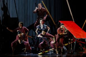 A large group of performers in red and brown costumes pose onstage holding black and red fans, with dramatic shadows cast on tall black curtains behind them and angular red wooden structures surrounding the ensemble.