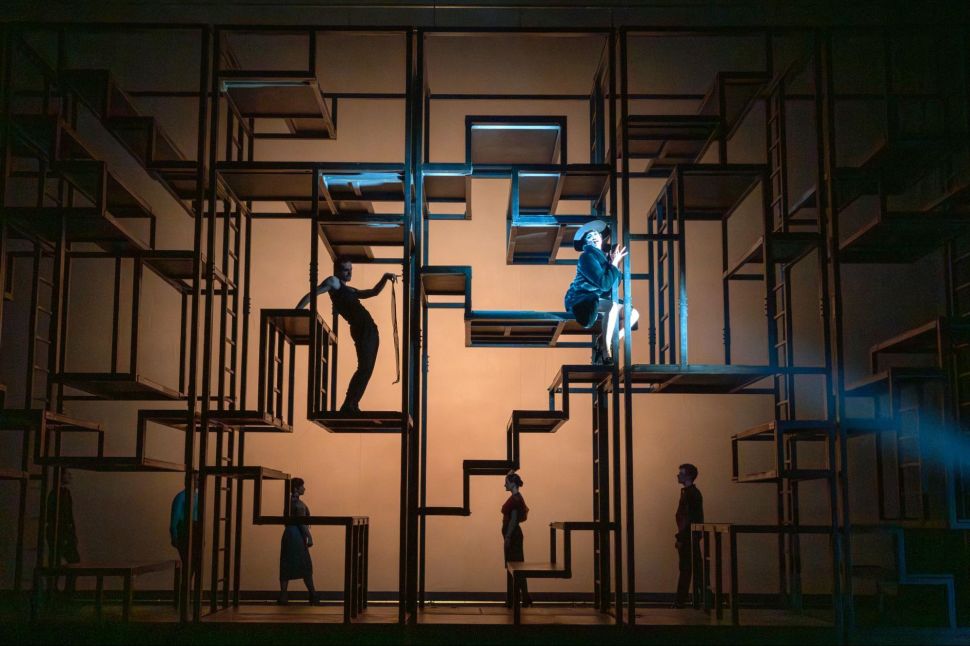 A wide shot of a theatrical set shows multiple actors standing or climbing within a grid-like structure of stacked metal rectangles, lit in silhouette and blue light against a flat beige backdrop.