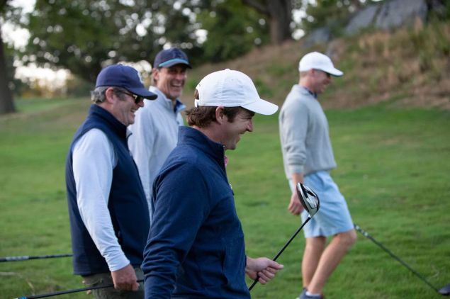 Four men in golf attire smiling and walking on a grassy fairway, holding clubs during a casual round of golf.