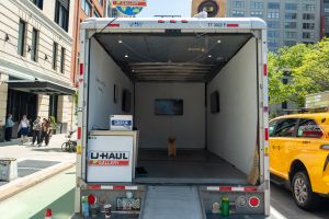 The open back of a U-Haul truck reveals a minimalist mobile gallery setup with white walls, three video screens mounted inside, and a single wooden bench on a rug.