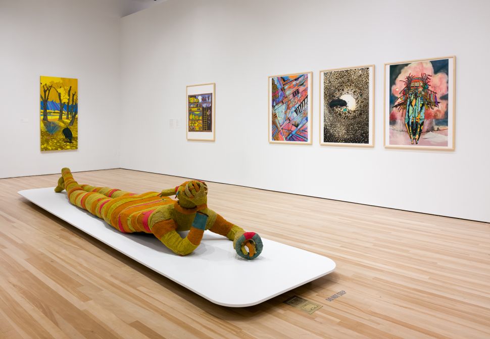 A brightly lit museum gallery features a life-sized, multicolored striped sculpture of a reclining human figure with its head resting on its hands, positioned on a low white platform in the center of the room; behind it, six framed artworks hang on the white walls, including abstract, narrative, and surreal images in vibrant colors, suggesting a group exhibition that blends sculpture and mixed media works.