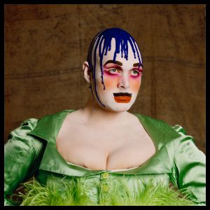 Leigh Bowery wears a vivid green satin dress with feather trim, dramatic clown-like makeup featuring blue paint dripping down a bald head, exaggerated eyebrows, bright pink eyeshadow, and bold red lips, embodying Bowery’s signature performance-art look.