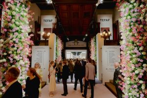 A floral-lined entrance at the Park Avenue Armory leads into the Wade Thompson Drill Hall during TEFAF New York, with visitors walking down a central corridor flanked by booths. The space is softly lit, and the left side of the frame is filled with a vibrant wall of flowers in shades of purple, pink, red and white.