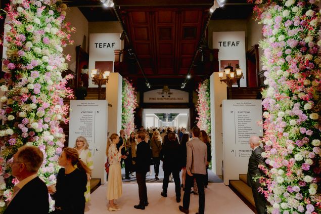 A floral-lined entrance at the Park Avenue Armory leads into the Wade Thompson Drill Hall during TEFAF New York, with visitors walking down a central corridor flanked by booths. The space is softly lit, and the left side of the frame is filled with a vibrant wall of flowers in shades of purple, pink, red and white.