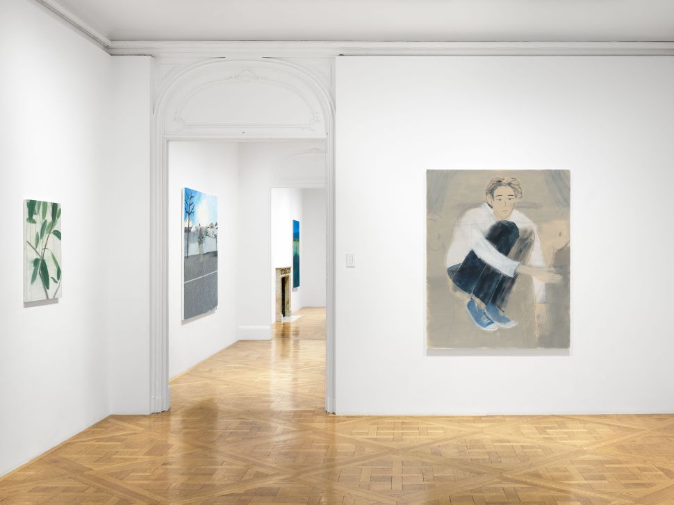 A hallway-like gallery space featuring multiple paintings. In the foreground, a large painting depicts a crouching figure in muted blues and grays. To the left, a botanical study leans toward minimalism. Further down the corridor, additional works line the walls, including a large-scale cityscape in icy blue tones.