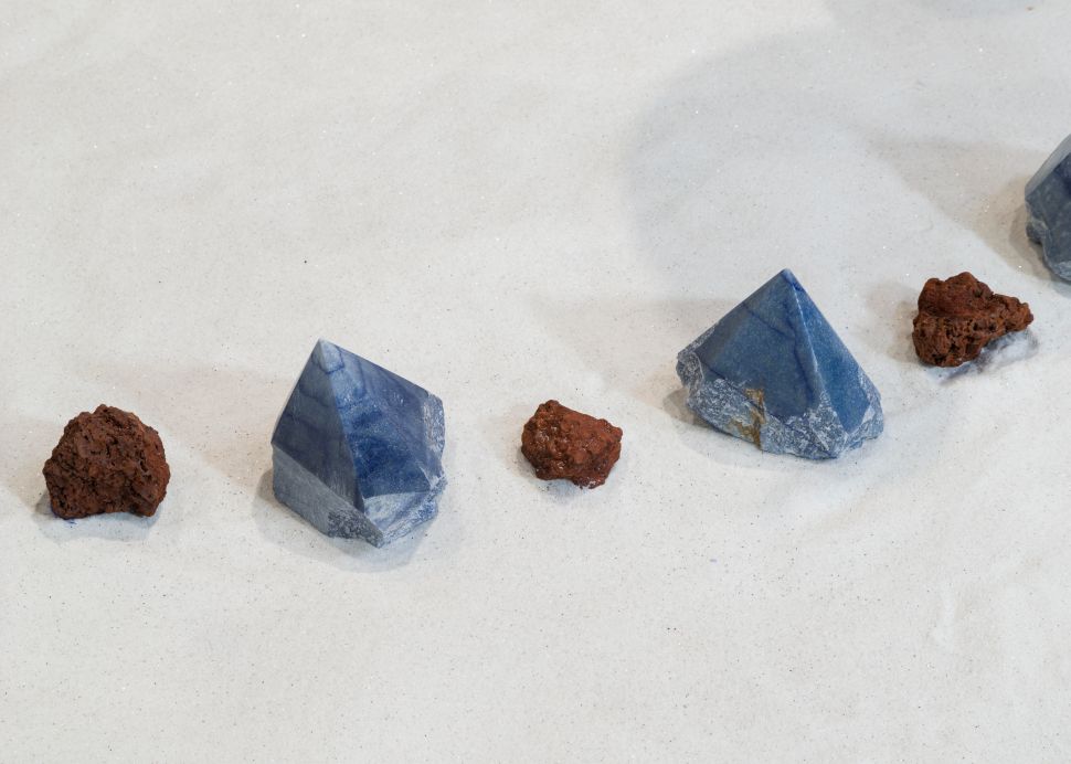 Close-up of alternating blue crystals and red volcanic rocks arranged in a linear path on a white granular surface, emphasizing the material contrast and tactile quality of the installation.