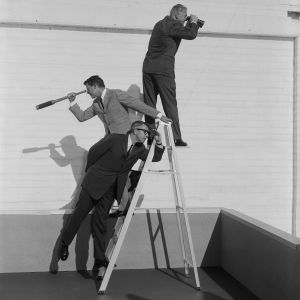 Three men in suits stand on and around a ladder, each using a different viewing tool—a telescope, binoculars, and spyglass—against a white brick wall.