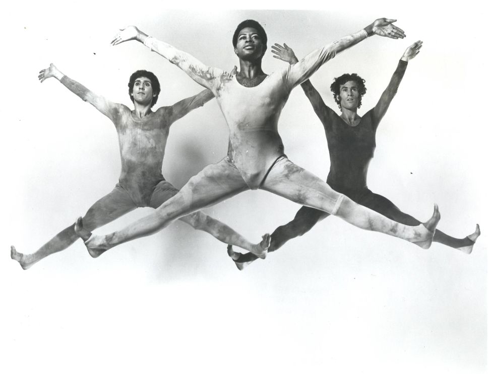 A black-and-white photograph shows three male dancers in midair performing identical grand jeté leaps with arms and legs fully extended in a line across the frame.