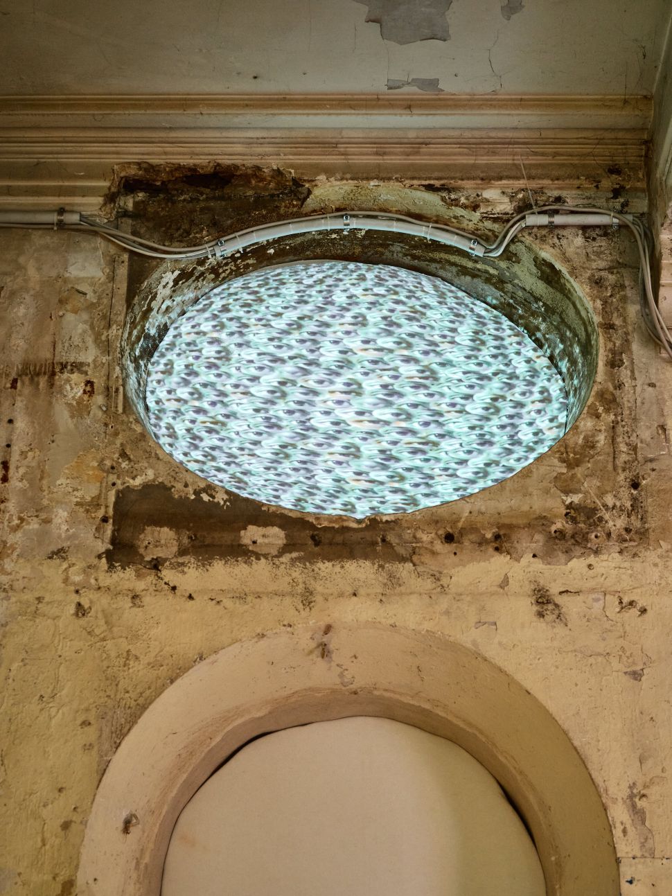 A circular projection of swirling water is embedded into a deteriorating, oval-shaped niche in a crumbling plaster wall, with exposed wiring framing the installation—evoking a portal or surveillance lens within a decayed architectural space.