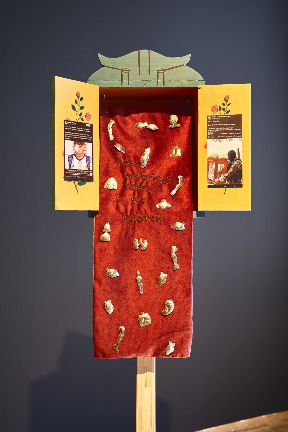 A handcrafted wooden retablo with open doors displays red velvet fabric adorned with tiny gold votive offerings and Spanish text reading “Las montañas saben lo que hicieron.”