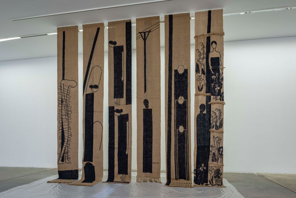 Tall vertical textile strips printed with black minimalist human forms and natural motifs hang in a white gallery space, evoking protest, mourning, and rural resistance.